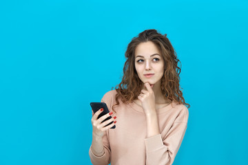 Fototapeta premium Portrait of a young beautiful woman wearing sweatshirt holding a mobile phone in her hand and looking at you thoughtfully over blue background