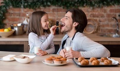 Caring little girl feeding her father with cookies in kitchen