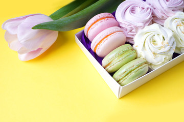 Box with macaroon and marshmallow cakes on a yellow background. Tulip lying near the box with sweets. Copy space.
