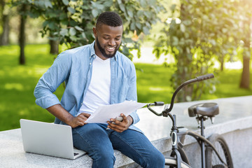 Happy Black Man With Laptop And Bike Checking Business Documentation Outdoors