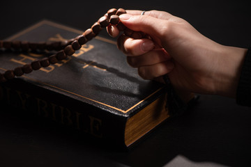 cropped view of woman holding rosary with cross near holy bible in dark with sunlight