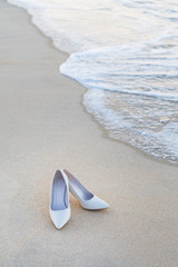 Fototapeta na wymiar Cream-colored bridal high heels Laying on the white sandy beach, luxurious, magnificent, the ocean waves blowing into the shore, along with the bubbles of free space, wedding celebrations.