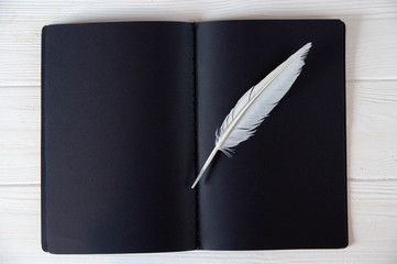 White feather on plack paper notebook on white wooden background, monochrome concept, writer work place