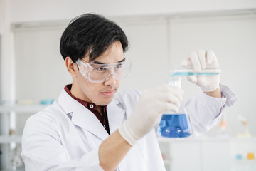 A male scientist with black hair wearing white coat and protective glassware pouring liquid solution from one test tube into a flask in a laboratory setting.