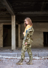Fototapeta na wymiar Young curly blond military woman, wearing ukrainian military uniform, posing for picture. Three-quarter portrait of soldier in front of ruined abandoned building, construction site.