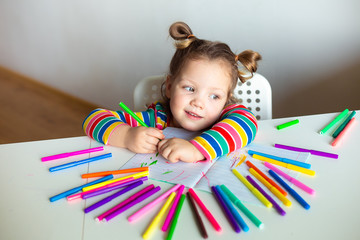 Little girl, a 3 year old girl, with a ponytail hairstyle in a multi-colored colorful striped jacket on a light background at the table draws multicolored markers and smiles
