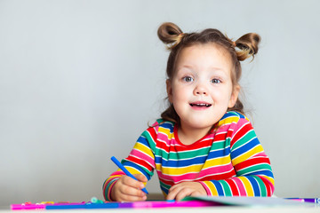 Little girl, a 3 year old girl, with a ponytail hairstyle in a multi-colored colorful striped jacket on a light background at the table draws multicolored markers and smiles