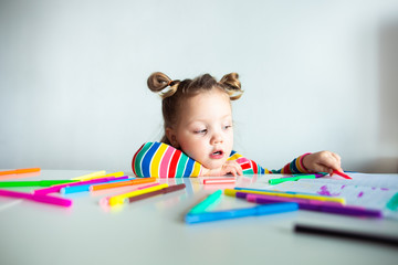 Little girl, a 3 year old girl, with a ponytail hairstyle in a multi-colored colorful striped jacket on a light background at the table draws multicolored markers and smiles