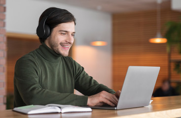 Young positive man in wireless headphones studying online on laptop