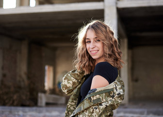 Young curly blond military woman, wearing ukrainian army military uniform and black t-shirt, posing. Three-quarter portrait of female soldier in front of ruined abandoned building,construction site.