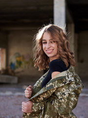 Fototapeta premium Young curly blond military woman, wearing ukrainian army military uniform and black t-shirt, posing. Three-quarter portrait of female soldier in front of ruined abandoned building,construction site.