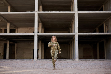 Young curly blond military woman, wearing ukrainian army military uniform and black t-shirt. Full-length portrait of female soldier standing in front of ruined abandoned building,construction site.