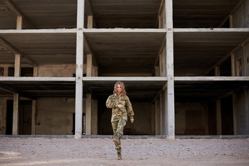 Young curly blond military woman, wearing ukrainian army military uniform and black t-shirt. Full-length portrait of female soldier standing in front of ruined abandoned building,construction site.