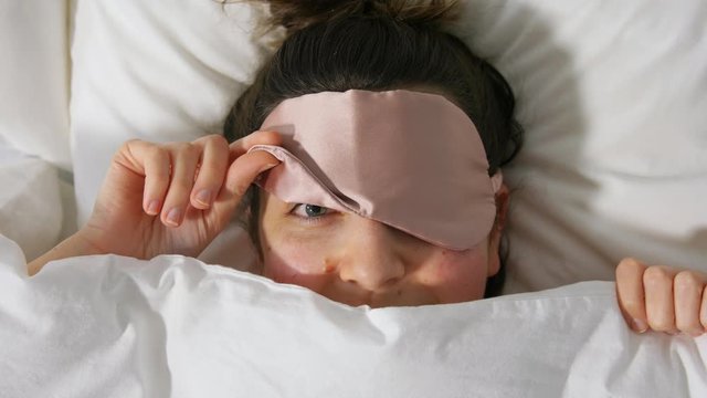people, bedtime and rest concept - happy woman in bed with eye sleeping mask peeking from under blanket