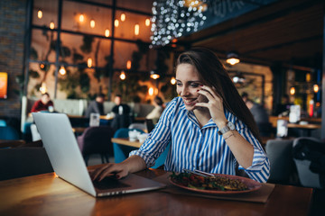 woman using mobile phone and laptop in restaurant while having lunch
