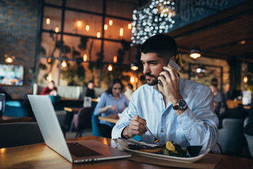 man using mobile phone and laptop in restaurant while having lunch