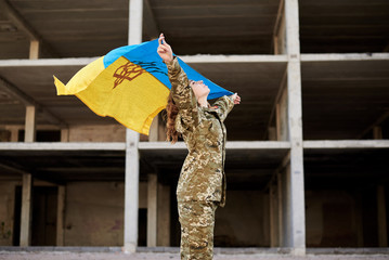 Young curly blond military woman, wearing ukrainian army uniform, holding ukrainian blue and yellow flag. Full-length portrait of female soldier in front of ruined abandoned building.