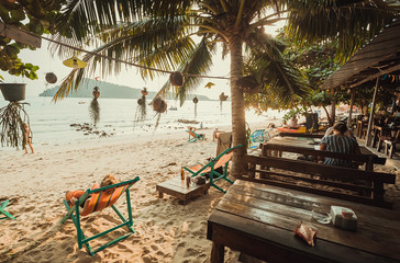 Beach restaurant with relaxing people, lounges on sand under palm trees and sea view. Happy sunny...