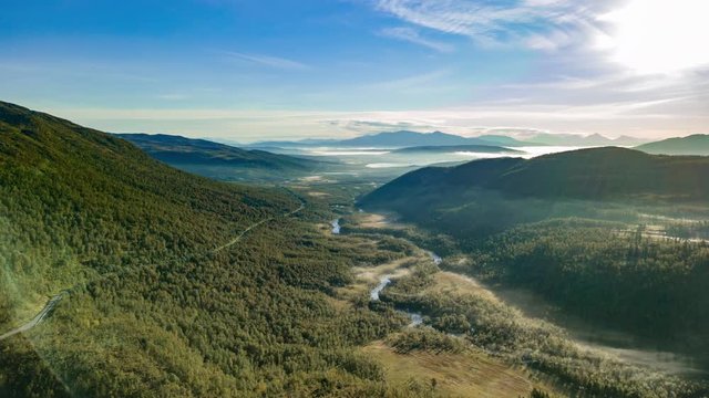 Magnificent view of the lush green valley of the Svanelva river. Blue sky above, thin, white, foglike clouds floating above the valley. Svanelva river meandering through the forest.