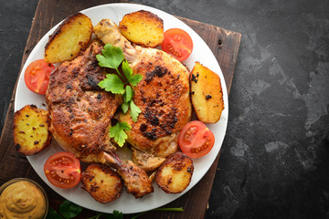 Fried chicken thigh, baked chicken leg. Top view. Dark background. Plate on a wooden board. Baked potatoes, tomatoes and chicken, a delicious lunch.