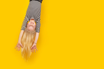 Excited crazy little blonde girl hanging happy upside down hands up over isolated yellow studio...