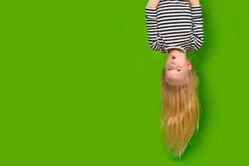 Surprised shocked little blonde girl hanging happy upside down over isolated green studio...