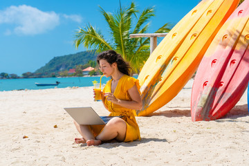 Girl in a yellow dress on a tropical sandy beach works on a laptop near kayaks and drinks fresh mango. Remote work, successful freelance. Works on vacation.