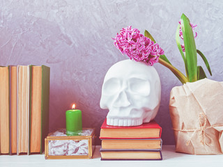Hyacinth flower wrapped in craft paper. Nearby is a figure in the form of a human skull on a stack of books, a burning candle, a glass casket and books. Gray concrete background. White wooden table.