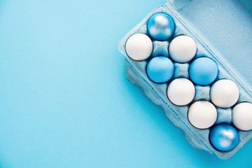 Close-up view of blue painted easter eggs and raw chicken eggs in egg box on  light blue background 