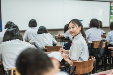 A pretty Asian female student in white uniform is turning her face and smiling among her friends while studying in the classroom.