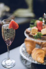 A close up of a strawberry on a champagne glass with croissants strawberries and cake on a tiered cake stand in the background