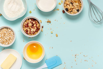 Ingredients for baking oat cookies with nuts and candied fruits on a blue background, top view, flat lay. Flour, butter, egg, oatmeal, sugar and nuts in bowls on the table, copy space
