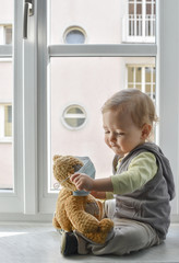 Child in home quarantine playing at the window with his sick teddy bear wearing a medical mask...