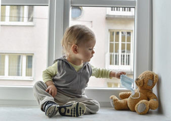 Child in home quarantine at the window putting a medical mask on his sick teddy bear, for...