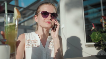 Fototapeta premium Slender Young Adult Woman with Ponytail and Sunglasses is Talking by Phone and Drinking Ice Tea Cocktail with Straw in the Terrace of Cafe or Restaurant in Summer