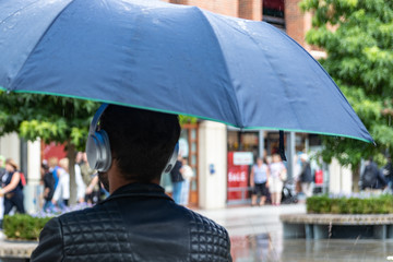 a man wearing headphones taking cover from rain under an umbrella at a shopping centre