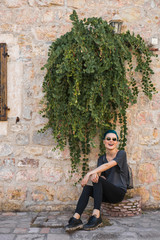 Fototapeta na wymiar Young girl in jeans and black sunglasses sits and laughs near a stone wall under an eucalyptus bush