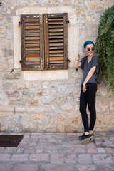 Fototapeta na wymiar Young girl in jeans and black sunglasses stands near a stone wall with wooden shutters
