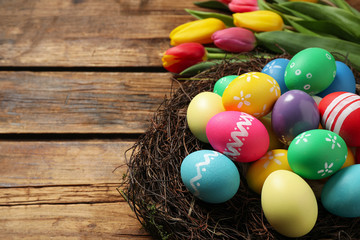 Colorful Easter eggs in decorative nest and tulips on wooden background, closeup. Space for text