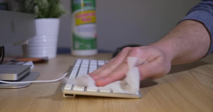 A slow dolly closeup view of a man cleaning his office computer's keyboard with a disinfectant wipe. Disease or flu prevention concept.  	