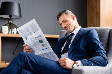 smiling businessman in suit sitting in armchair and reading newspaper with business lettering