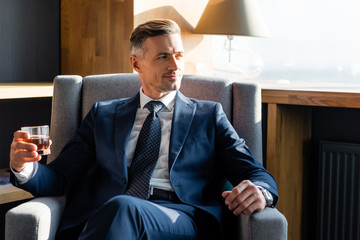 smiling businessman in suit sitting in armchair and holding glass