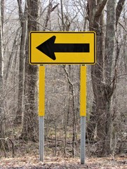 View of a yellow direction traffic sign on the side of a country road with woods in the background 