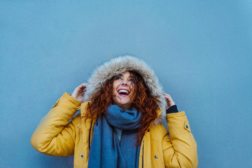 Cheerful woman looking up with a hood winter yellow coat about to rain. Female isolated on blue background with copy space. Bright complementary colors. Travel and winter holidays concept. 