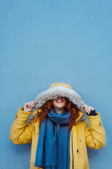 Cheerful woman looking up with a hood winter yellow coat protection from rain. Female isolated on blue background with copy space. Bright complementary colors. Travel and winter holidays concept. 