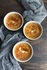 Homemade creme brulee - traditional French vanilla cream dessert with caramel decor, caramelised brown sugar on top in black baking dishes, wooden table, grey textile. Top view, flat lay, copy space.