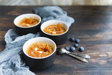 Homemade creme brulee - traditional french vanilla cream dessert with caramel decor, caramelised brown sugar on top in black baking dishes, wooden  table, grey textile and spoons. Copy space.