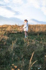 Beautiful hippie girl with flower bouquet, wearing white clothes, dress and jacket in boho style, hat and cowboy boots, walking in the summer field