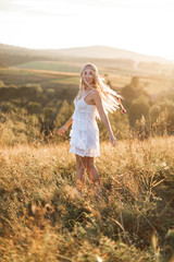 Beautiful young boho styled woman in white dress and with feathers in hair walking at summer field, sunset