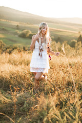 Fototapeta na wymiar Young blond beautiful hippie girl in white dress and feather hair accessories on summer field and sunset background
